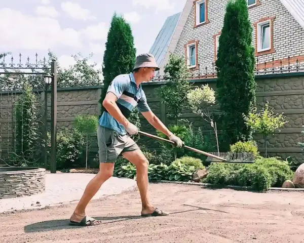 Un homme en chapeau et short utilise un râteau pour faire une place de parking dans son jardin. Le soleil brille sur la pelouse.