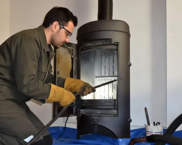 Un homme en tenue de travail effectue le ramonage d'un poêle à bois, ganté et muni d'outils.