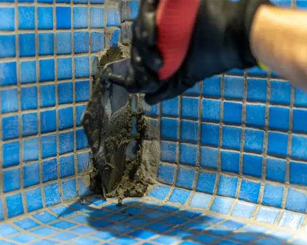 Application de mortier pour l'étanchéité d'une piscine en béton. Un ouvrier en gant noir et rouge applique le produit avec une spatule sur les carreaux bleus.