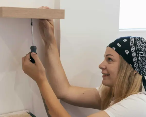 Une femme souriante, bandeau sur la tête, utilise un tournevis pour fixer une étagère en bois. Elle sait comment percer droit pour un résultat parfait.