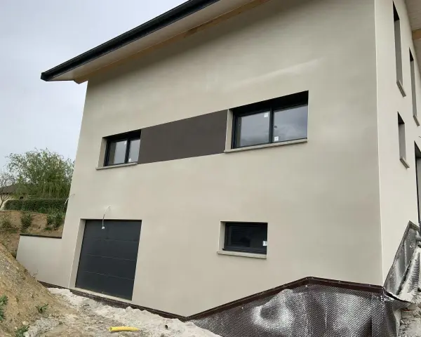 Maison moderne avec enduit neuf, une trace blanche discrète sur le crépi clair. Fenêtres noires et garage gris.