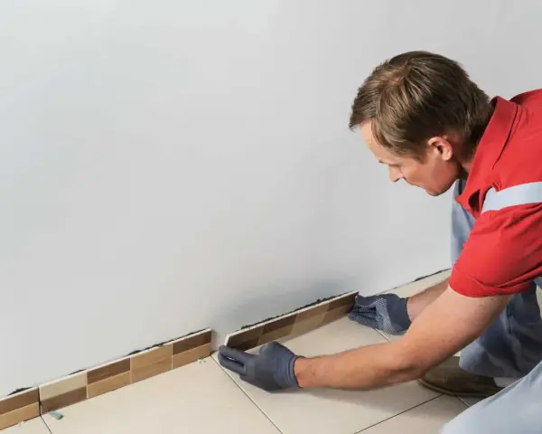 Un homme en gants gris est en train d'enlever la plinthe de carrelage le long d'un mur blanc.