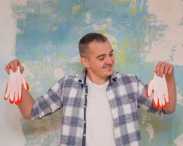 Homme souriant tenant des gants blancs à bouts orange devant un mur texturé. Il pense au prix peinture m2 au black pour son projet.
