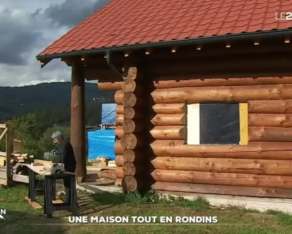 Une maison en rondins de bois en construction, avec un homme travaillant à proximité. Le toit est rouge et le paysage est montagneux.