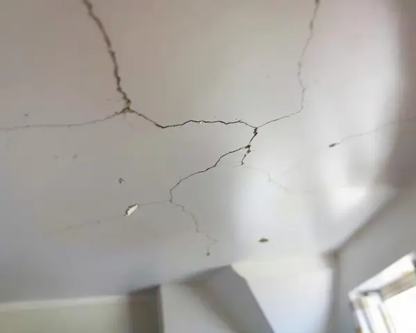 Un plafond qui s'effrite, révélant des fissures sombres et des morceaux de plâtre tombés. La lumière douce éclaire les imperfections.