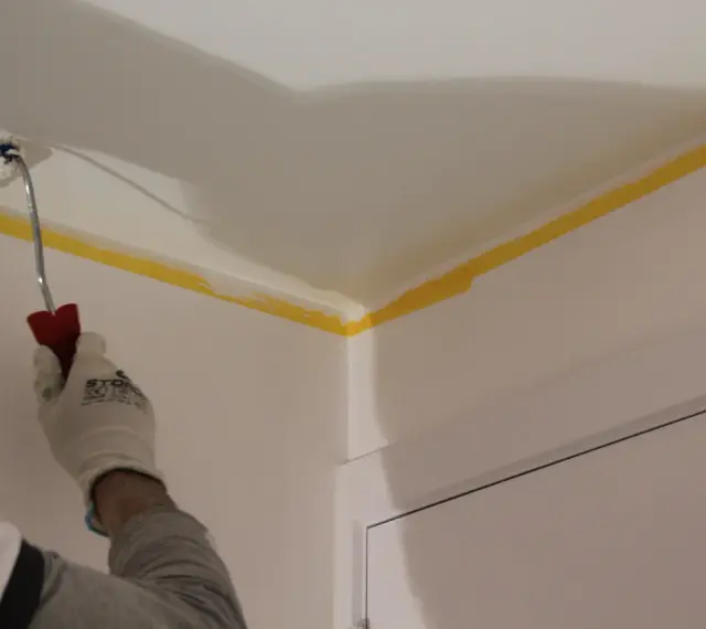 Maintenant, vous savez comment peindre un plafond : une main gantée utilise un rouleau pour peindre le coin d'un plafond, protégé par du ruban de masquage jaune.