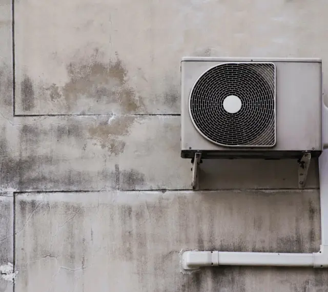 Unité extérieure d'une pompe à chaleur air air fixée sur un mur en béton fissuré et taché.