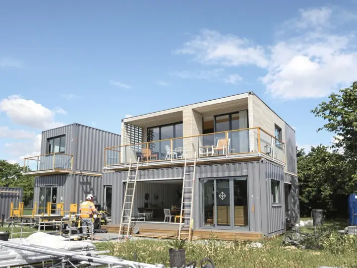 Maison en container prix : construction moderne à deux étages avec balcons, fenêtres panoramiques et un ouvrier en gilet de sécurité.