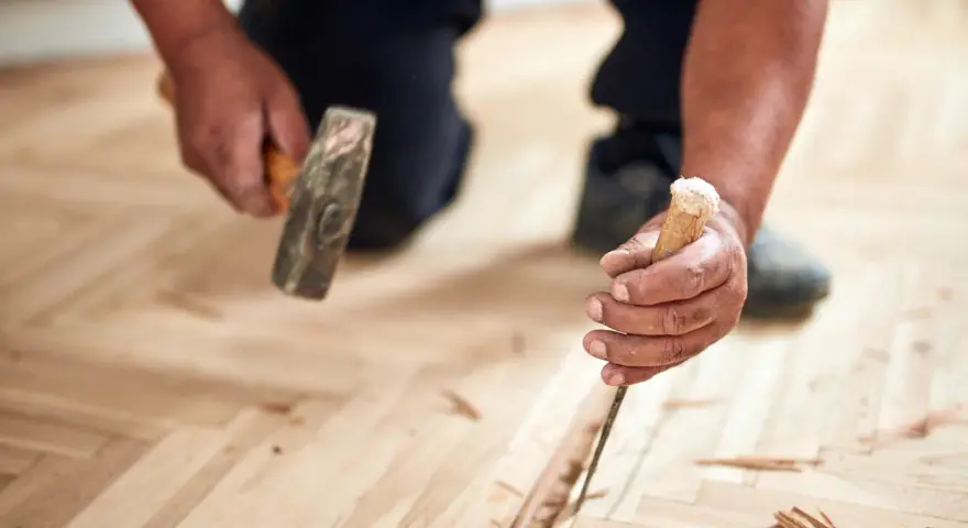 Un artisan utilise un ciseau et un marteau pour changer une lame de parquet flottant endommagée.