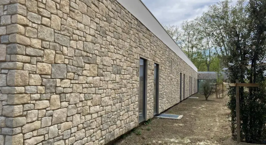 Un mur extérieur en pierres naturelles irrégulières, avec des fenêtres modernes et un toit blanc. Le sol est en terre battue.