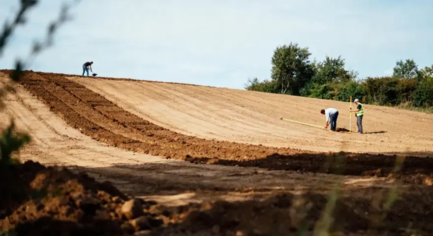 Des ouvriers travaillent sur un terrain en pente, utilisant des outils pour aplanir le sol. L'un mesure avec un mètre, l'autre pelle.