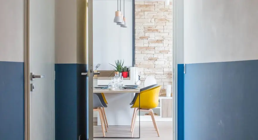 Peinture bicolore sur les murs d'un couloir et de portes, ouvrant sur une salle à manger moderne avec des chaises colorées.