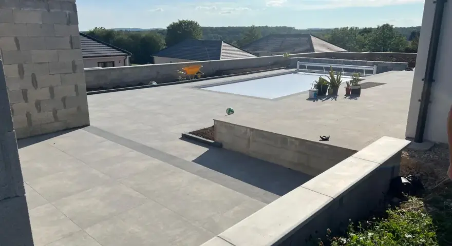 Vaste terrasse en béton gris avec piscine couverte, entourée de végétation et de maisons. Un chantier en cours.