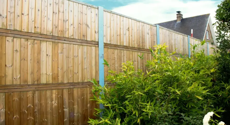 Clôture anti bruit en bois, haute et solide, protégeant un jardin verdoyant. Le ciel bleu et les nuages ajoutent une touche de sérénité.