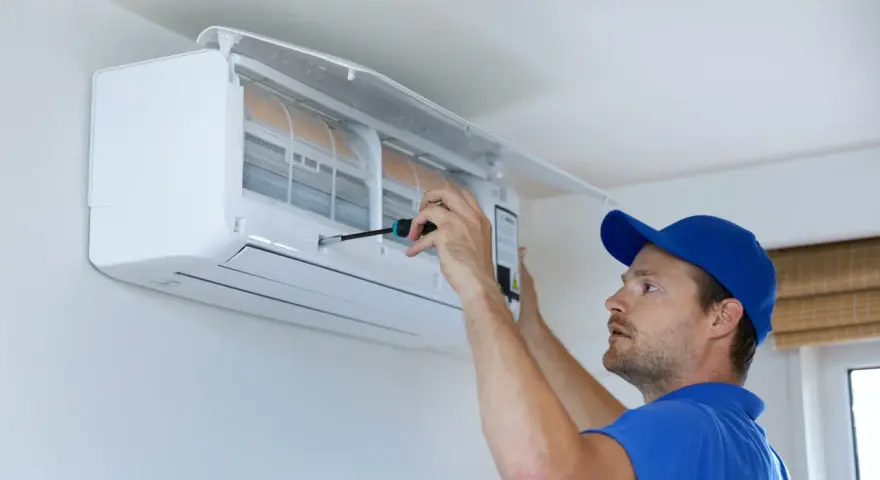 Un technicien en bleu utilise un tournevis pour ajuster un climatiseur mural, assurant le bon fonctionnement pour recharger clim maison.
