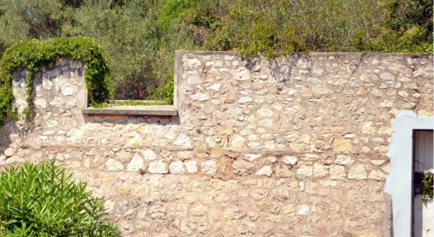 Un mur en pierre extérieur, avec des pierres irrégulières et du mortier, attend de restaurer son charme ancien. La végétation s'y accroche.