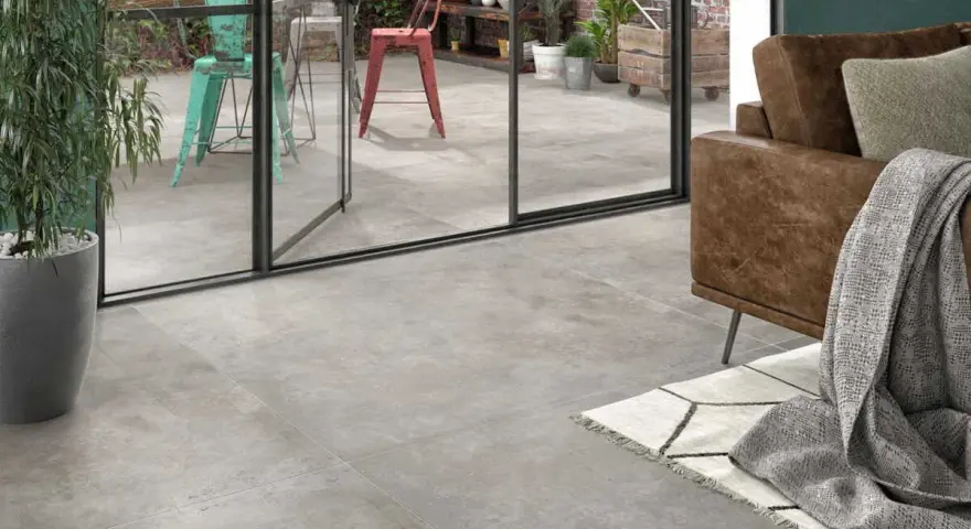 Un salon moderne avec un canapé en cuir marron et un plaid gris. Le sol est recouvert d'un carrelage usé que faire ? Une grande baie vitrée donne sur un patio avec des chaises colorées.