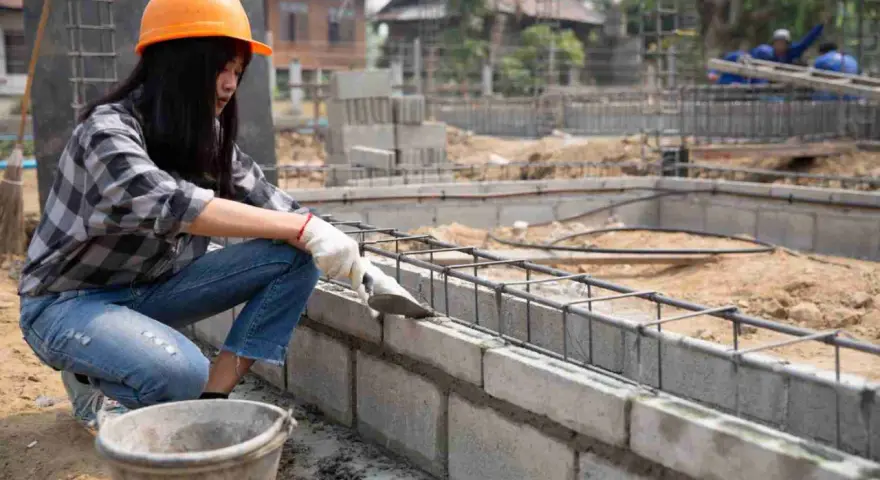 Une ouvrière en casque orange pose du ciment sur un mur en parpaing. Le chantier progresse.