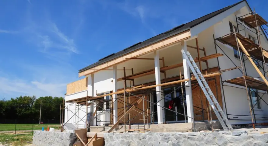 Une maison en construction, avec des échafaudages et une échelle, en cours d'agrandissement. Le ciel bleu est parsemé de nuages légers.