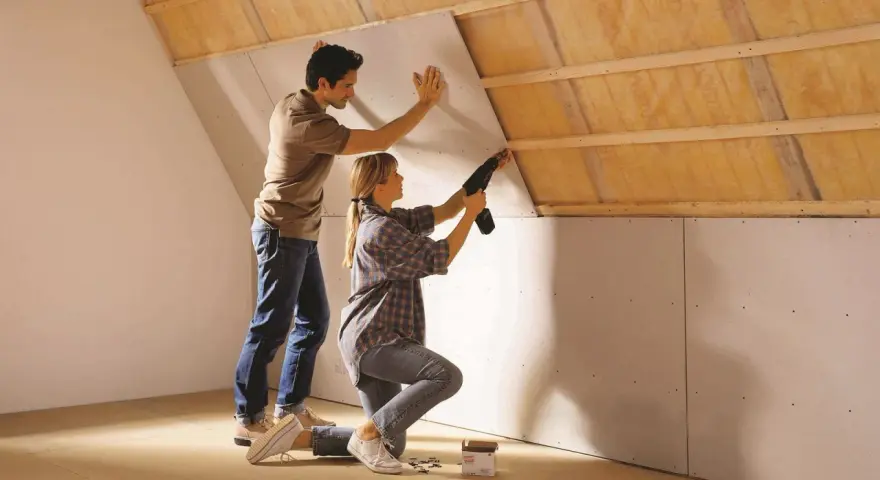 Un couple installe des plaques de plâtre sur un mur mansardé, préparant la surface avant un enduit à la chaux.