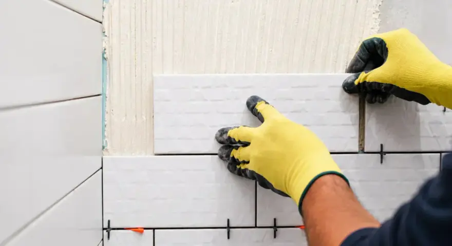Pose-t-on coller du carrelage avec du ciment ? Oui, un carreleur pose du carrelage blanc sur un mur avec des croisillons.