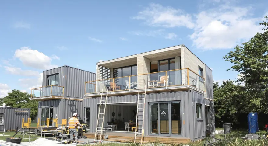 Maison en container prix : construction moderne à deux étages avec balcons, fenêtres panoramiques et un ouvrier en gilet de sécurité.
