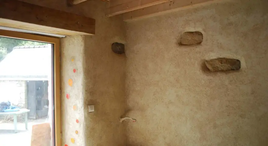 Un mur avec un enduit chaux finition texturé, intégrant des pierres apparentes et des touches de couleur. Le bois des poutres et du cadre de fenêtre apporte de la chaleur.
