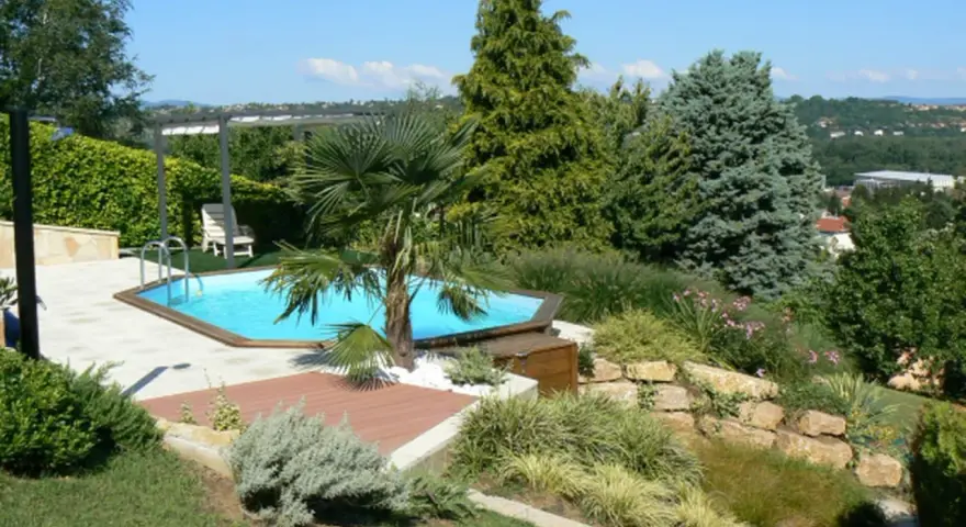 Piscine aux formes douces sur un terrain en pente, entourée de végétation luxuriante et d'une terrasse en bois.