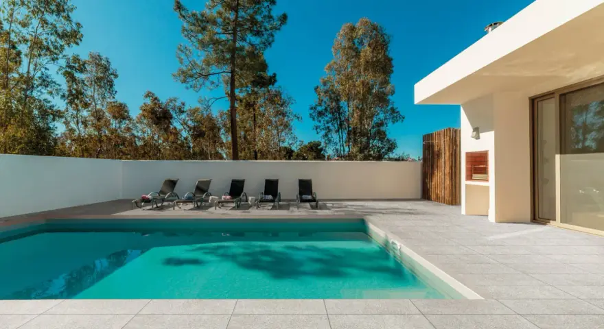 Une piscine moderne avec un sol en carrelage gris clair, des chaises longues et un barbecue extérieur sous un ciel bleu éclatant.