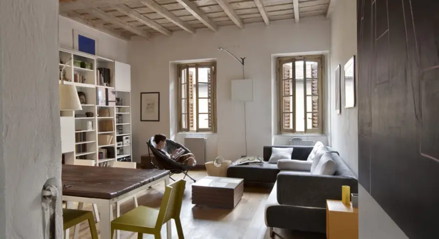 Salon avec poutres apparentes au plafond, une personne lisant dans un fauteuil, canapé, table et étagères.
