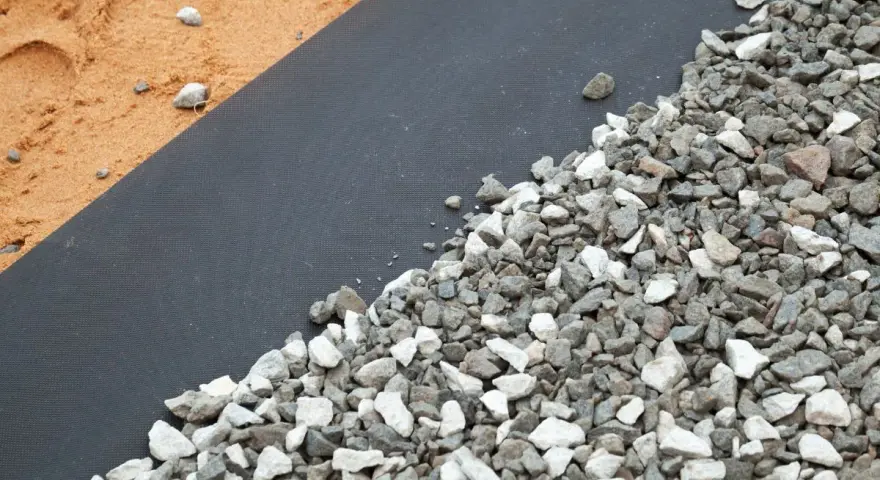 Gros plan sur un géotextile noir recouvert de gravier gris et blanc, à côté de sable. Idéal pour estimer le prix pose géotextile et gravier.