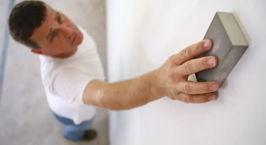 Un homme ponce un mur blanc avec une cale à poncer, préparant la surface avant la peinture.