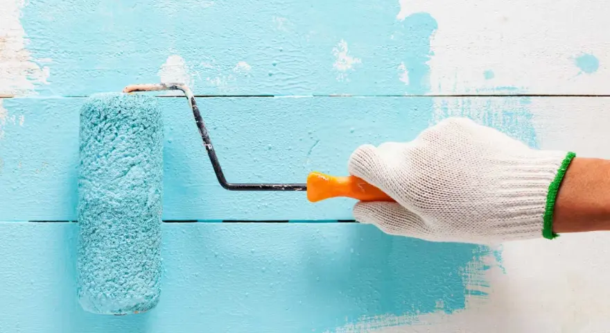Main tenant une main gantée, un rouleau applique une peinture bleu turquoise sur un mur en bois blanc.