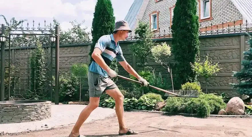 Un homme en chapeau et short utilise un râteau pour faire une place de parking dans son jardin. Le soleil brille sur la pelouse.