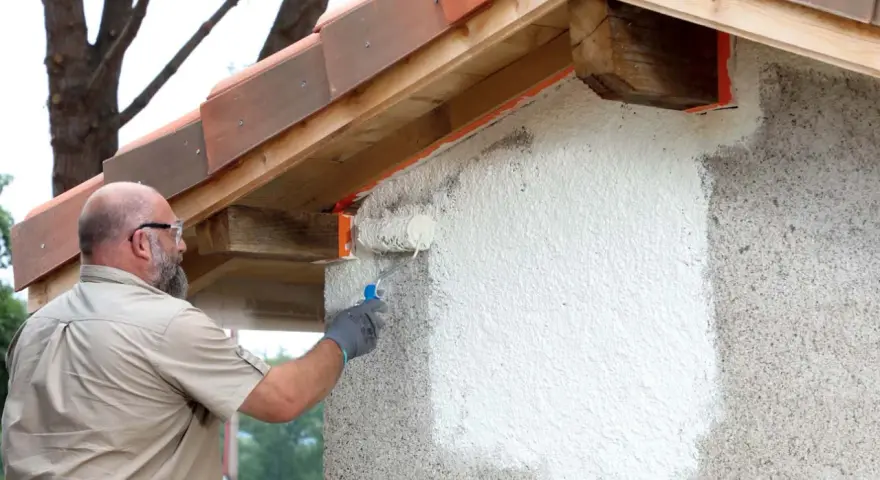 Un homme applique une peinture sur mur béton extérieur avec un rouleau. La rénovation progresse sous le toit de tuiles.