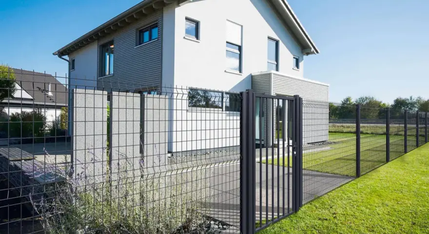 Une clôture privative en limite de propriété délimite un jardin moderne avec une maison blanche et un portillon.