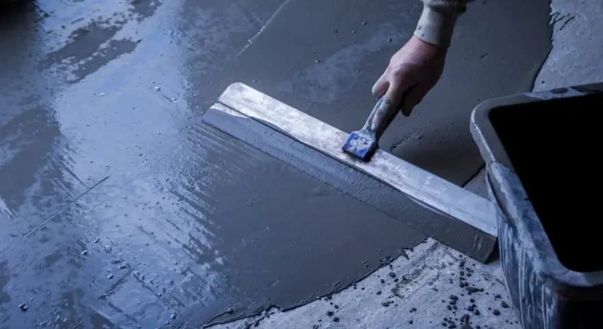 Main d'un ouvrier réalisant un ragréage de terrasse extérieur avec une lisseuse, assurant une pente pour l'évacuation de l'eau.