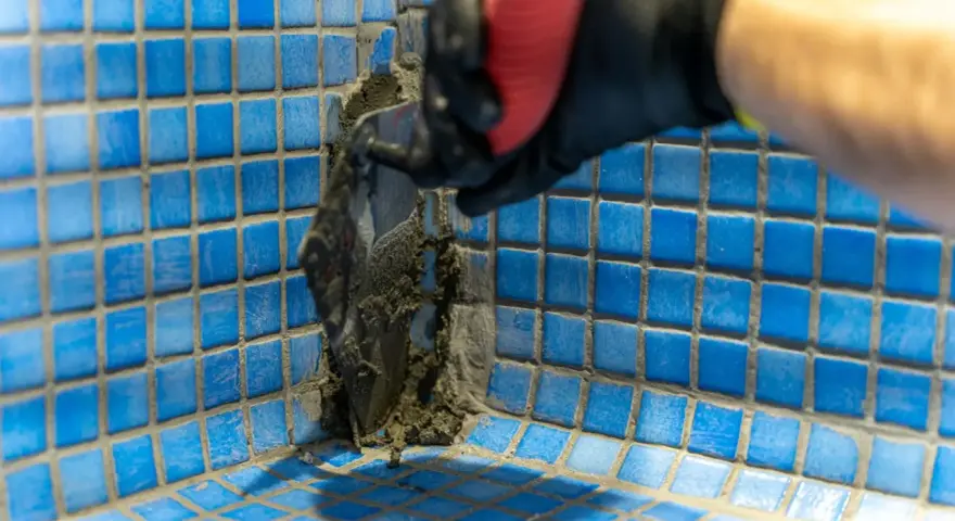 Application de mortier pour l'étanchéité d'une piscine en béton. Un ouvrier en gant noir et rouge applique le produit avec une spatule sur les carreaux bleus.