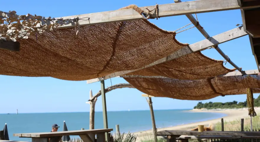 Installation d'une voile d'ombrage en toile de jute sur une structure en bois, offrant une vue sur la mer et la plage.