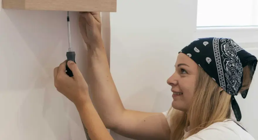 Une femme souriante, bandeau sur la tête, utilise un tournevis pour fixer une étagère en bois. Elle sait comment percer droit pour un résultat parfait.