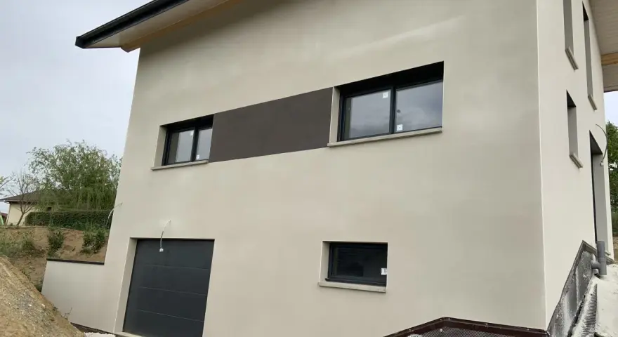 Maison moderne avec enduit neuf, une trace blanche discrète sur le crépi clair. Fenêtres noires et garage gris.