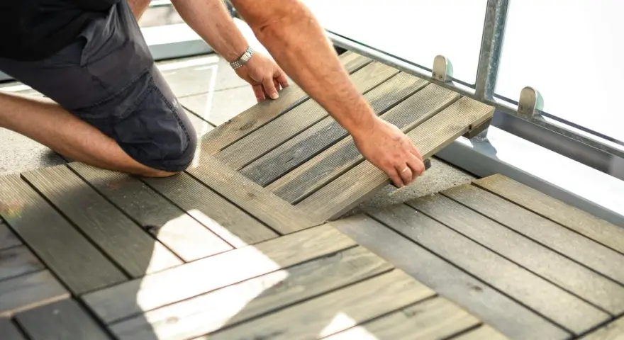 Un homme installe des dalles de terrasse en bois sur son balcon. Une belle idée sol balcon pour un espace extérieur accueillant.