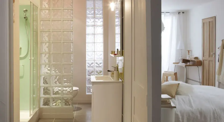 Salle de bain moderne avec douche, lavabo et toilettes, séparée de la chambre par un mur en brique de verre.