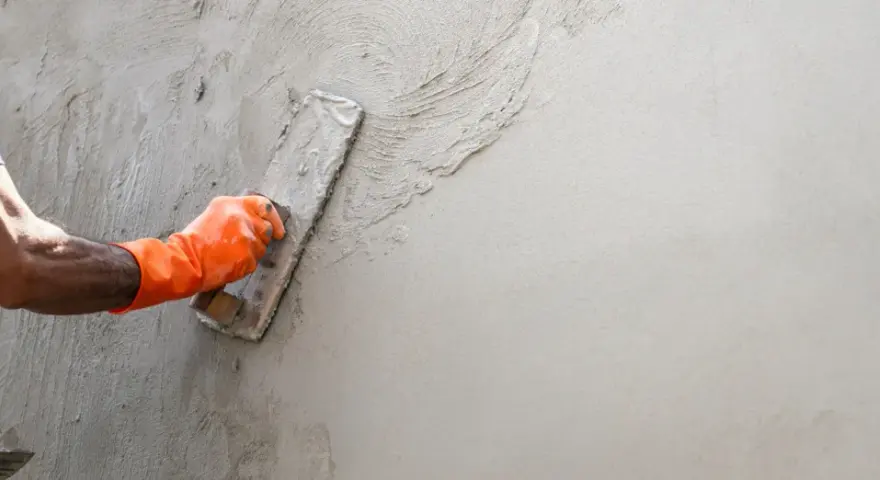 Main d'un ouvrier en gant orange appliquant de l'enduit sur un mur avec une truelle. Apprentissage de comment faire un enduit sur un mur.