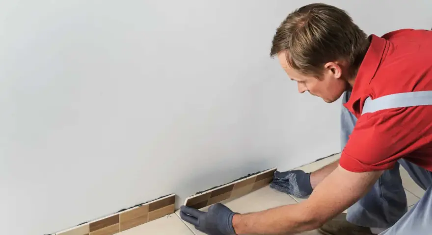 Un homme en gants gris est en train d'enlever la plinthe de carrelage le long d'un mur blanc.