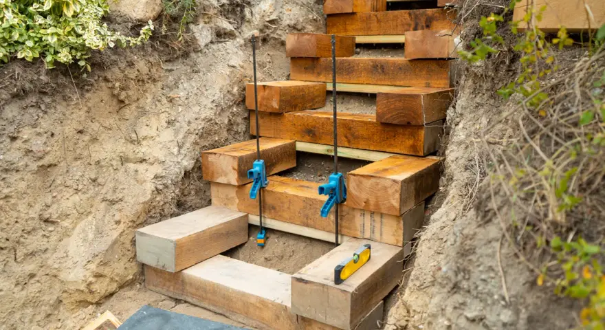 Construction d'un escalier traverse bois et gravier. Des poutres de bois sont empilées pour former des marches dans une tranchée.