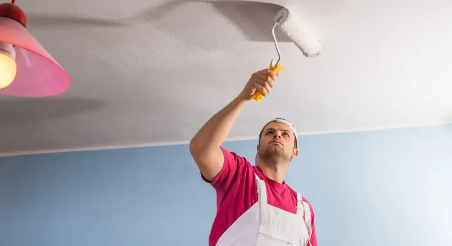 Un homme en salopette blanche et t-shirt rose utilise un rouleau pour peindre un plafond. Il est concentré sur sa tâche, montrant comment peindre un plafond.