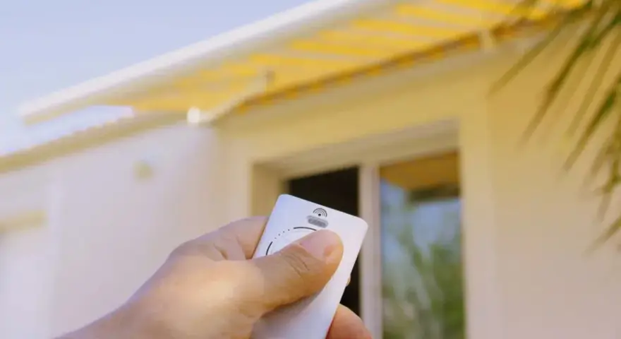 Main tenant une télécommande pour le réglage store banne manuel. Le store jaune est déployé sur une maison blanche.