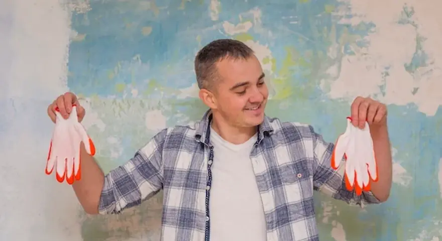 Homme souriant tenant des gants blancs à bouts orange devant un mur texturé. Il pense au prix peinture m2 au black pour son projet.