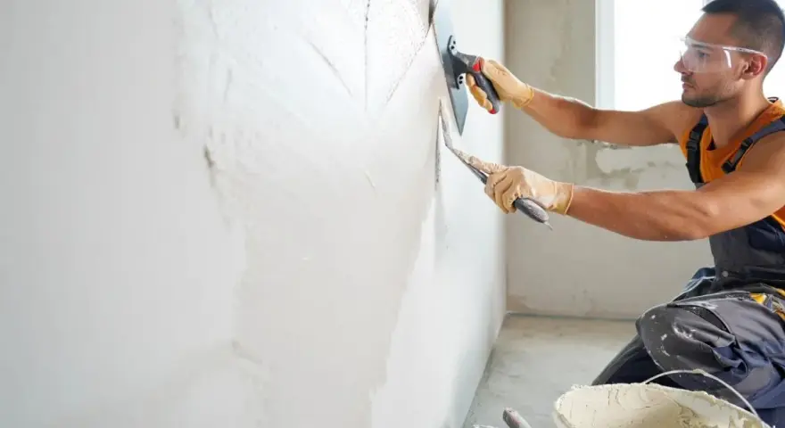 Un ouvrier applique un enduit sur un mur humide avec une truelle et une taloche. Le travail progresse, laissant une surface lisse.